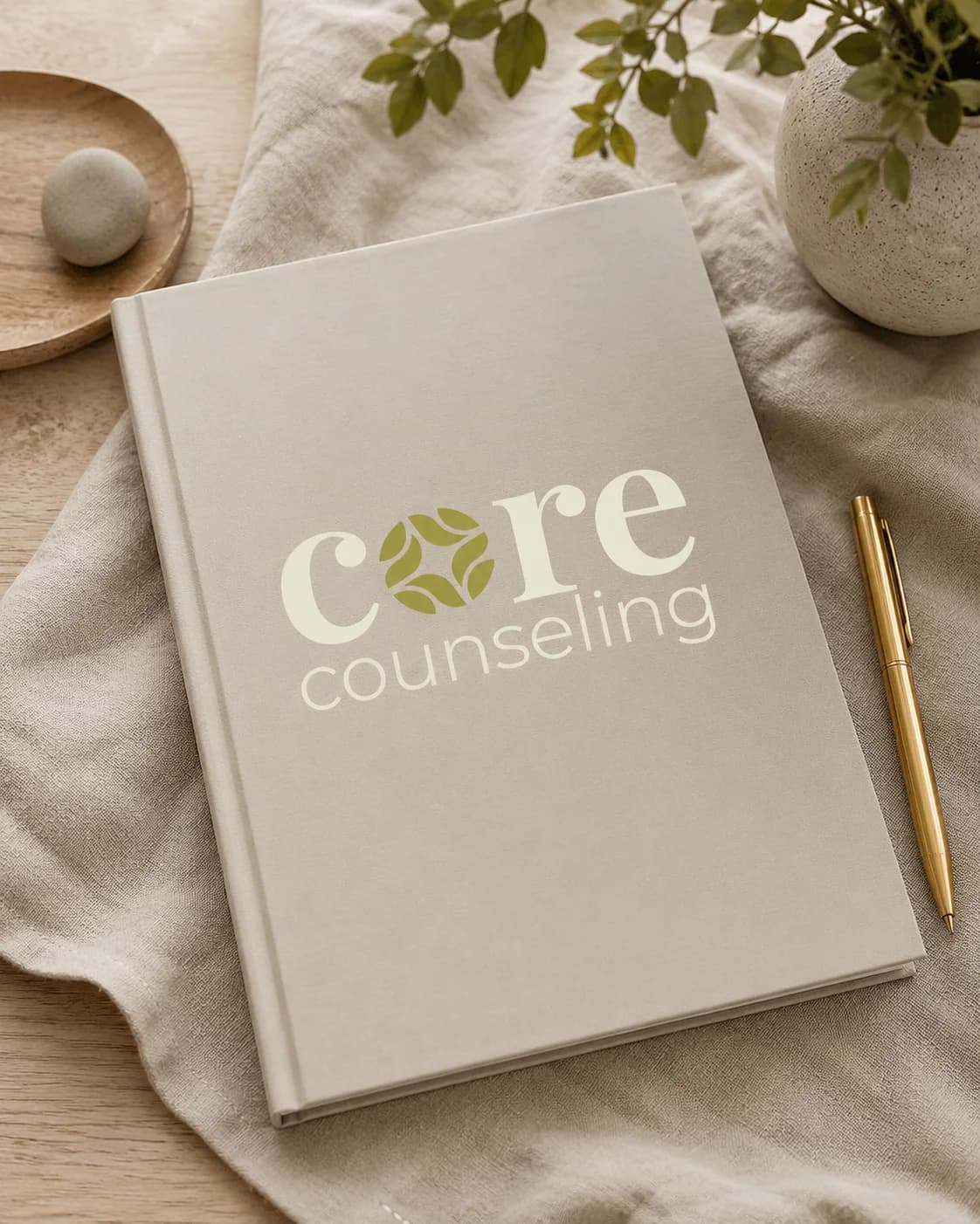 Linen-bound notebook with the Core Counseling logo in cream and olive, beside a gold pen and a sprig of greenery